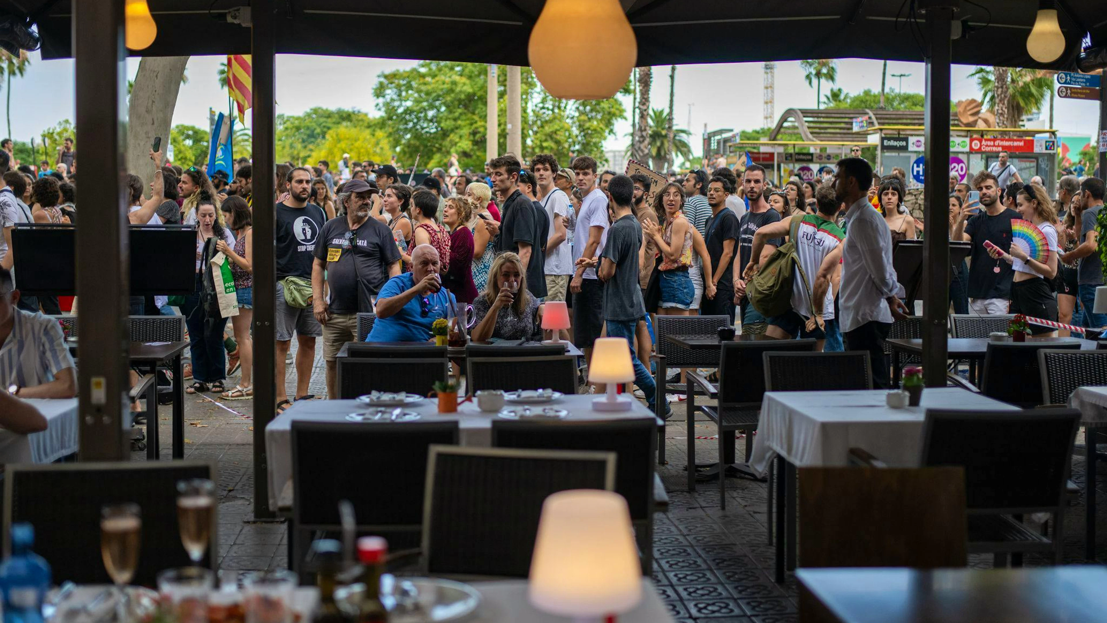 Ein Mann und eine Frau auf der Terrasse eines Restaurants in Barcelona lassen sich von vorbeiziehenden Demonstranten gegen den Massentourismus offenbar nicht aus der Ruhe bringen.