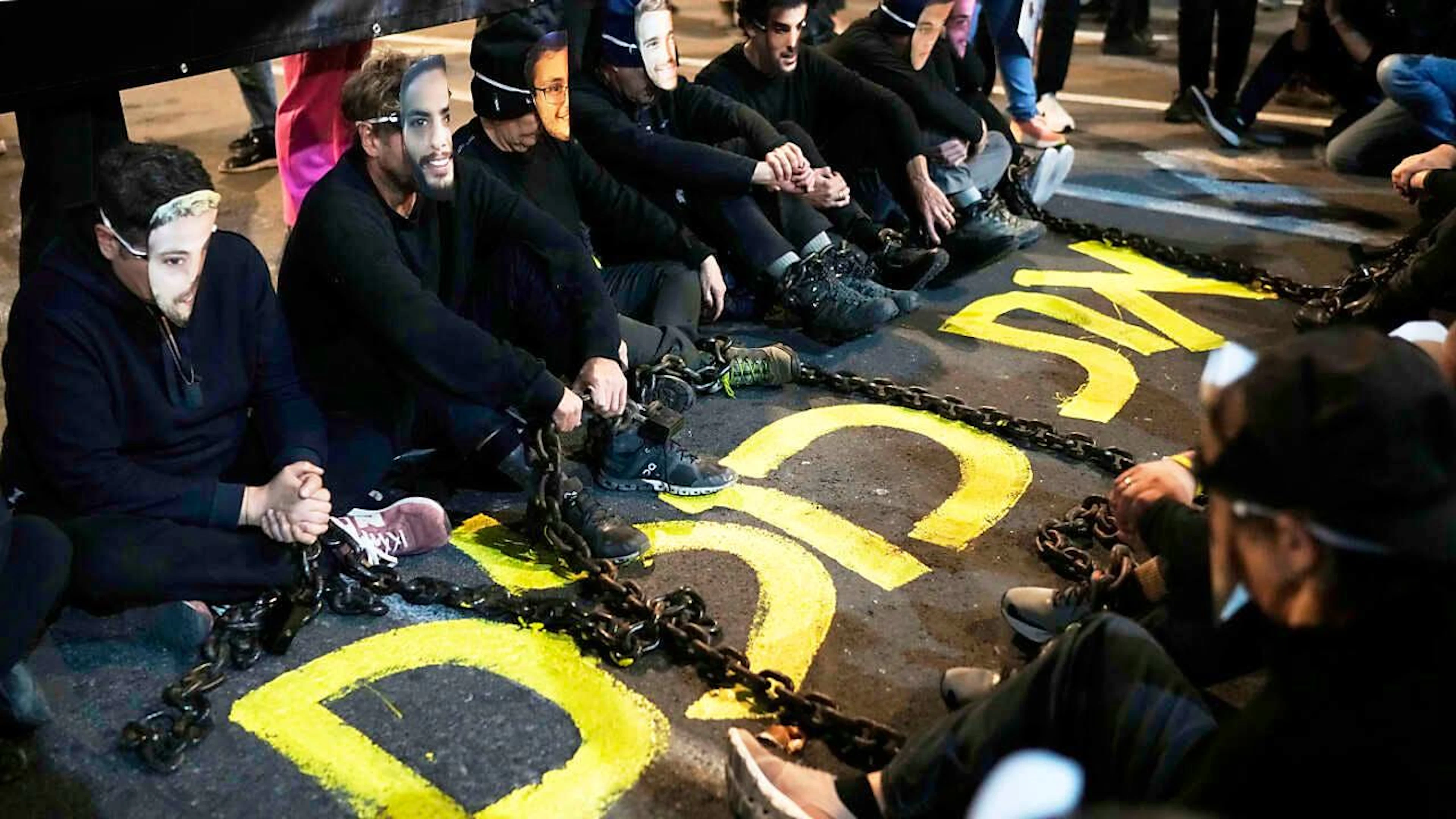 Aktivisten sitzen mit Eisenketten auf einer Straße in Tel Aviv und tragen Masken mit den Gesichtern von Geiseln, die von der Terrororganisation Hamas immer noch im Gazastreifen festgehalten werden. Foto: Leo Correa/AP/dpa