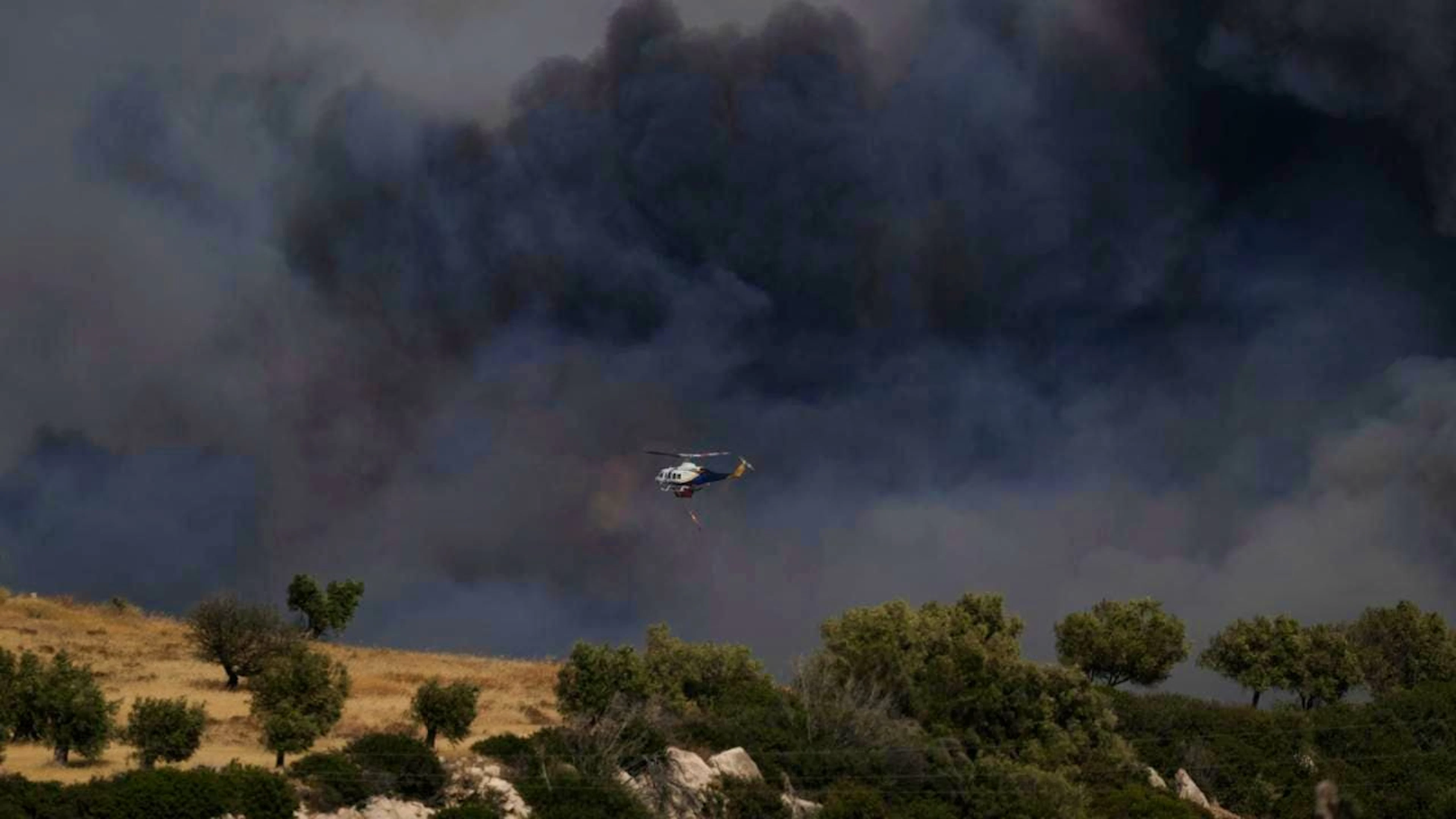 Ein Hubschrauber fliegt durch den Rauch eines Brandes in Keratea, einem Außenbezirk von Athen. Foto: Thanasis Stavrakis/AP/dpa