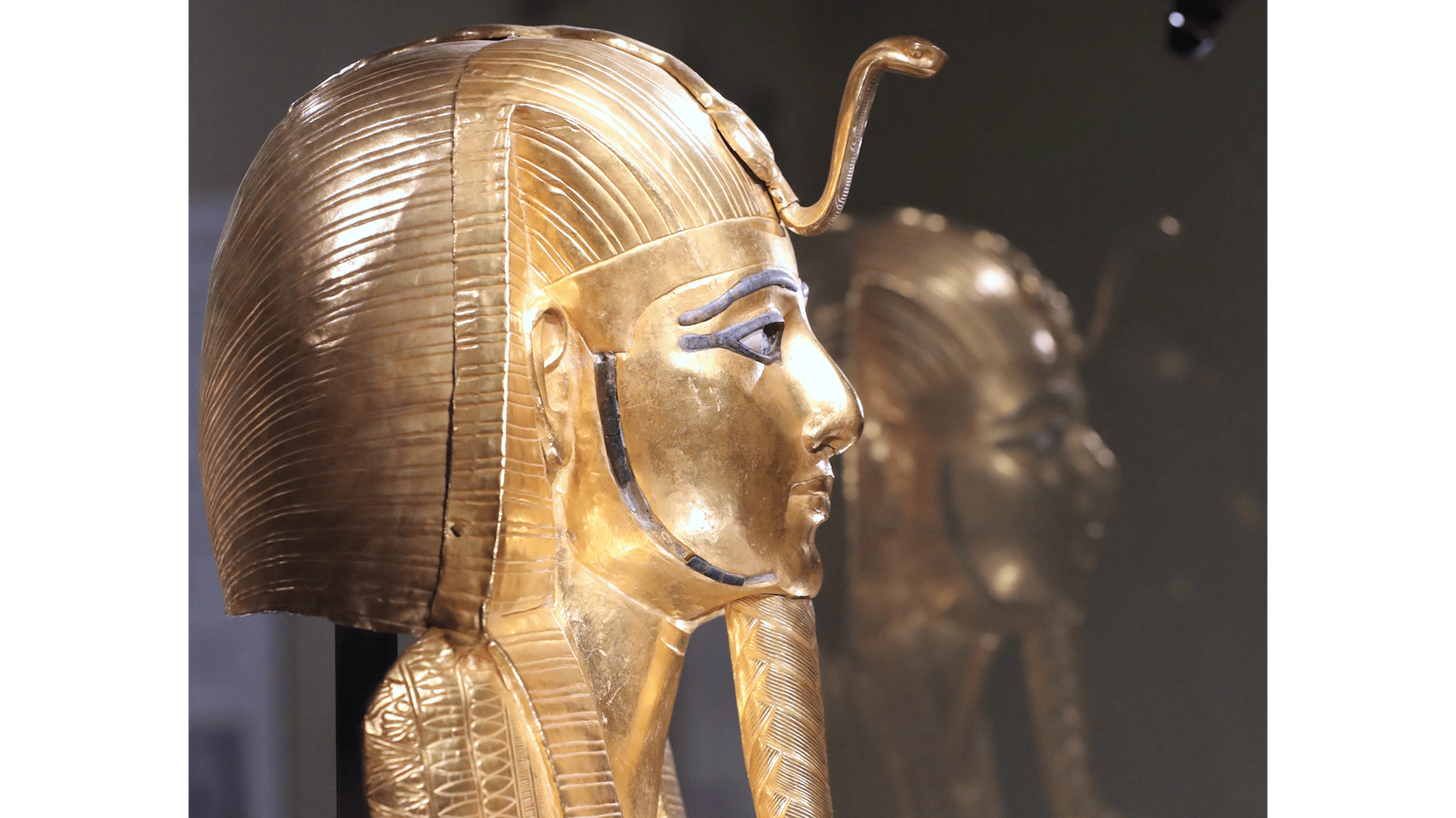 Die lebensgrosse Goldmaske des Psusennes I. mit ruhigen, idealisierten Gesichtszügen: Sie trägt das gestreifte Nemes-Kopftuch, auf der Stirn sitzt die Uräus-Kobra als königliches Schutzsymbol. Auffällig sind die dunkel konturierten Augen und Details in blau – sie entstehen durch Einlagen aus Lapislazuli und Glasfluss. Am Kinn sitzt der geflochtene Zeremonialbart, ein typisches Zeichen königlicher Würde.