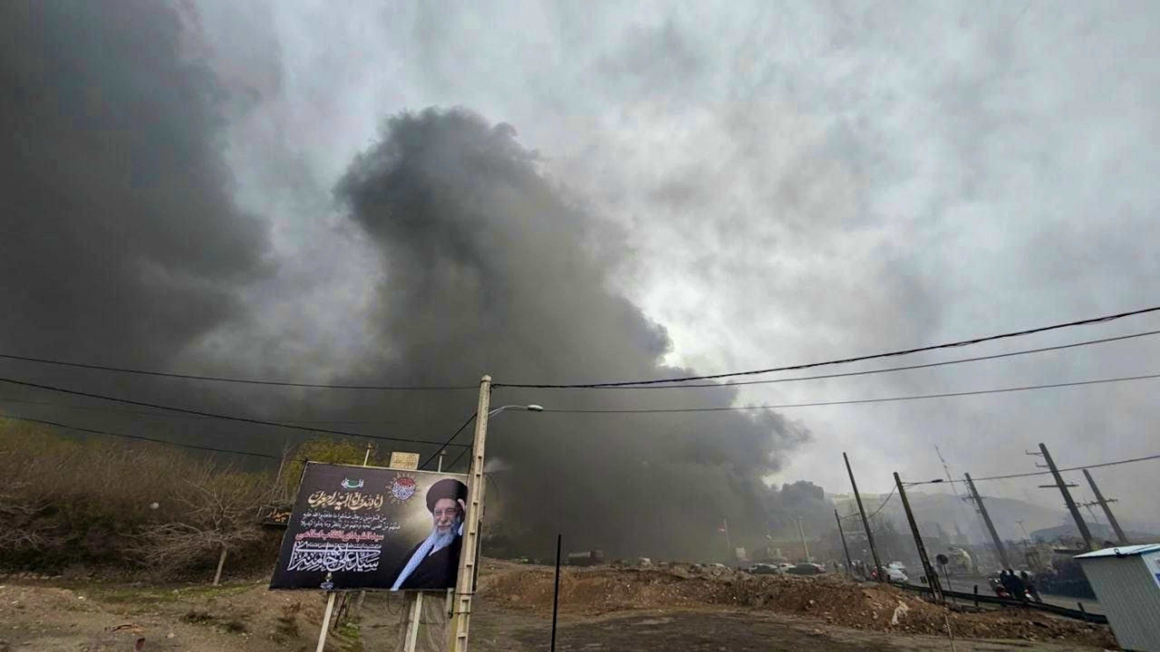 A thick cloud of smoke resulting from a US-Israeli attack on an oil facility late Saturday is seen in the cloudy sky behind a billboard showing the late Iranian Supreme Leader Khamenei, in Tehran. Photo: Vahid Salemi/AP /dpa