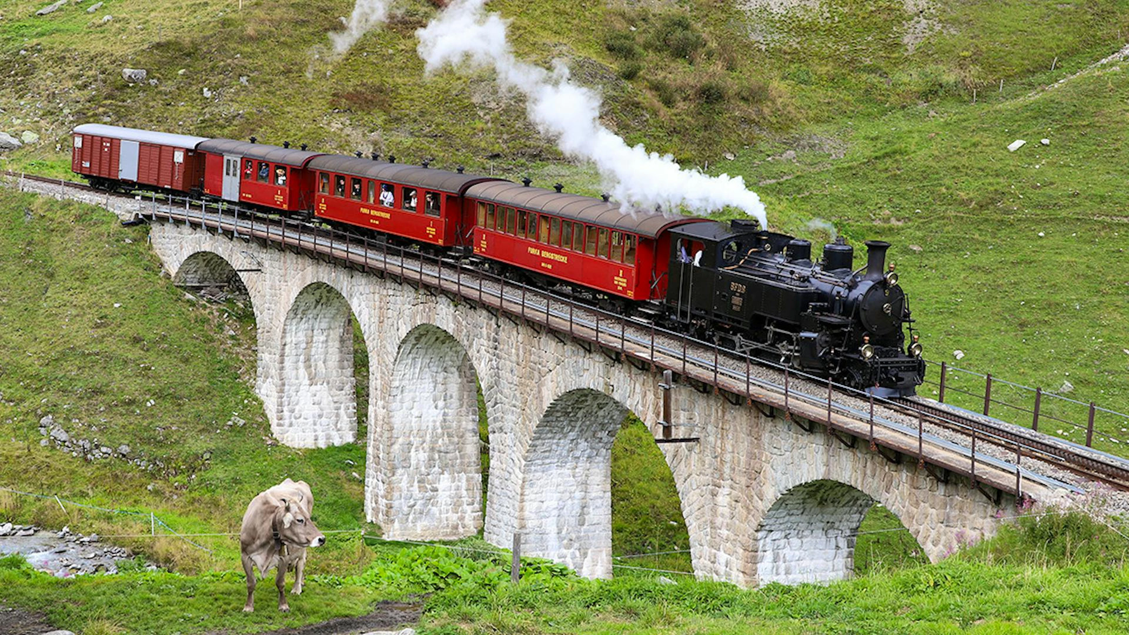 Die historische Dampfbahn Furka-Bergstrecke überquert mit ihren roten Wagen einen Viadukt in den Urner Alpen – ein Stück lebendige Schweizer Bahngeschichte.