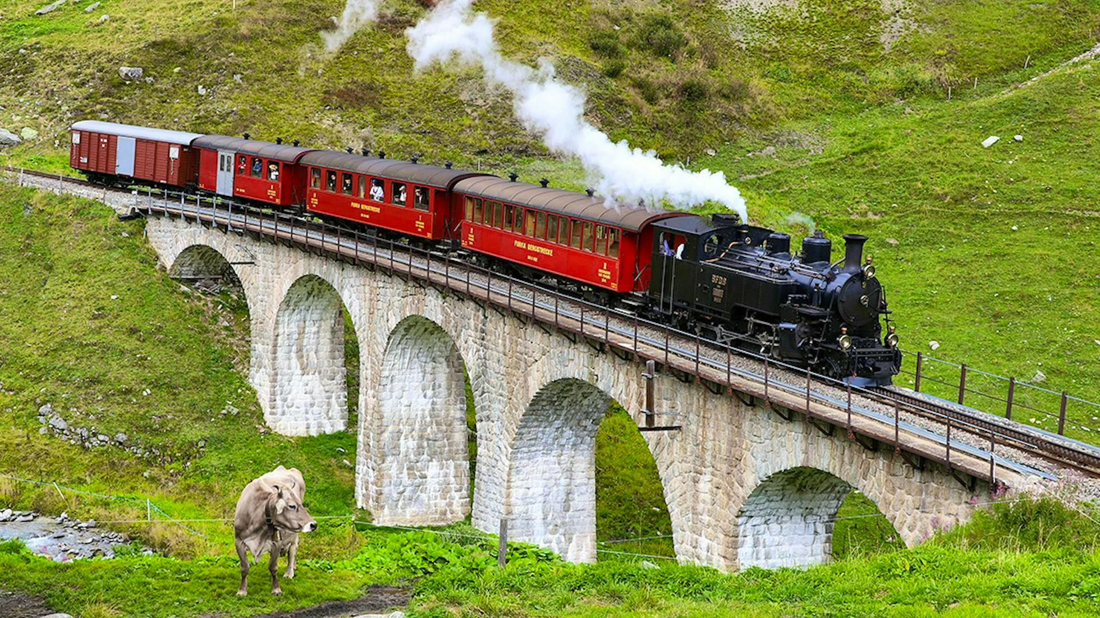 The historic Furka-Bergstrecke steam railroad crosses a viaduct in the Uri Alps with its red carriages - a piece of living Swiss railroad history.
