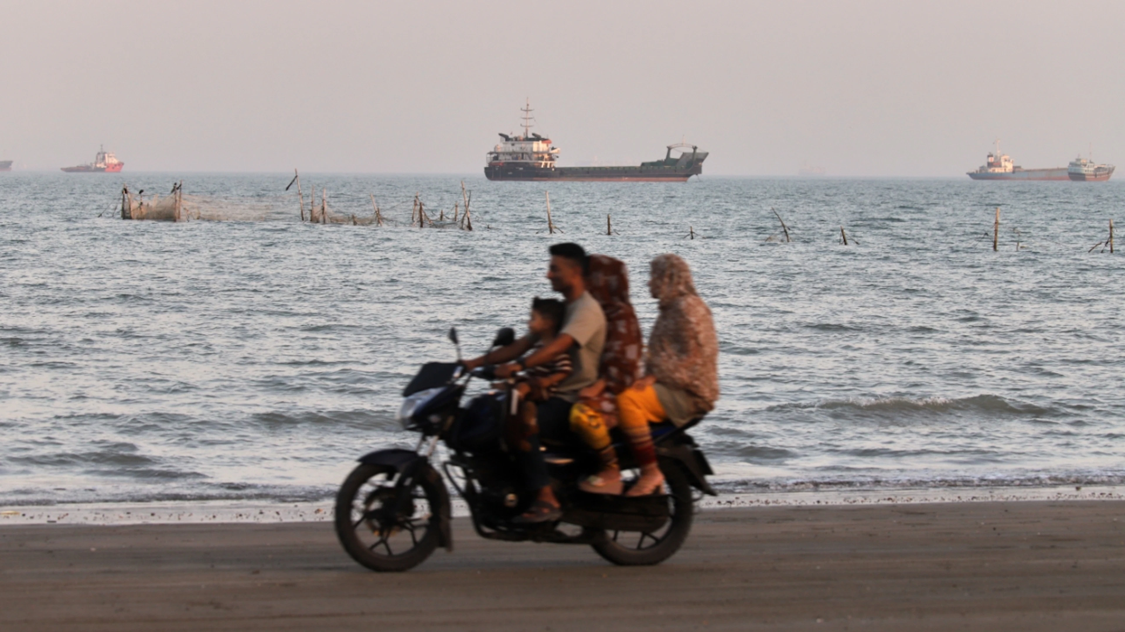 ARCHIV – Fracht- und Serviceschiffe säumen den Horizont, während Menschen auf einem Motorrad über den Strand am Ufer der Strasse von Hormus fahren. Foto: Amirhosein Khorgooi/ISNA/dpa/Symbolbild
