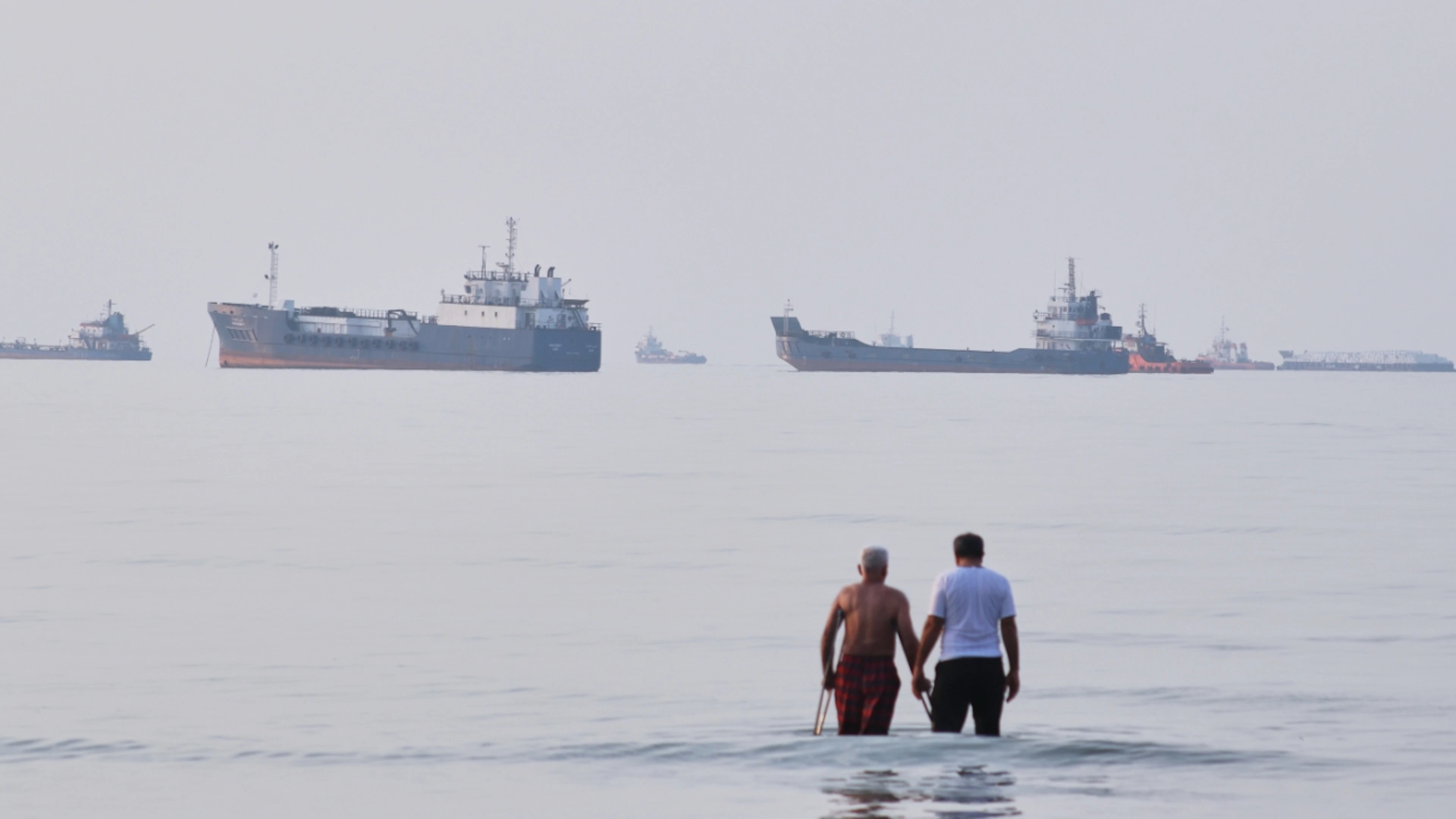 Two men wade through the water in the Strait of Hormuz, while ships lie at anchor in the background. Photo: Razieh Poudat/ISNA via AP/dpa