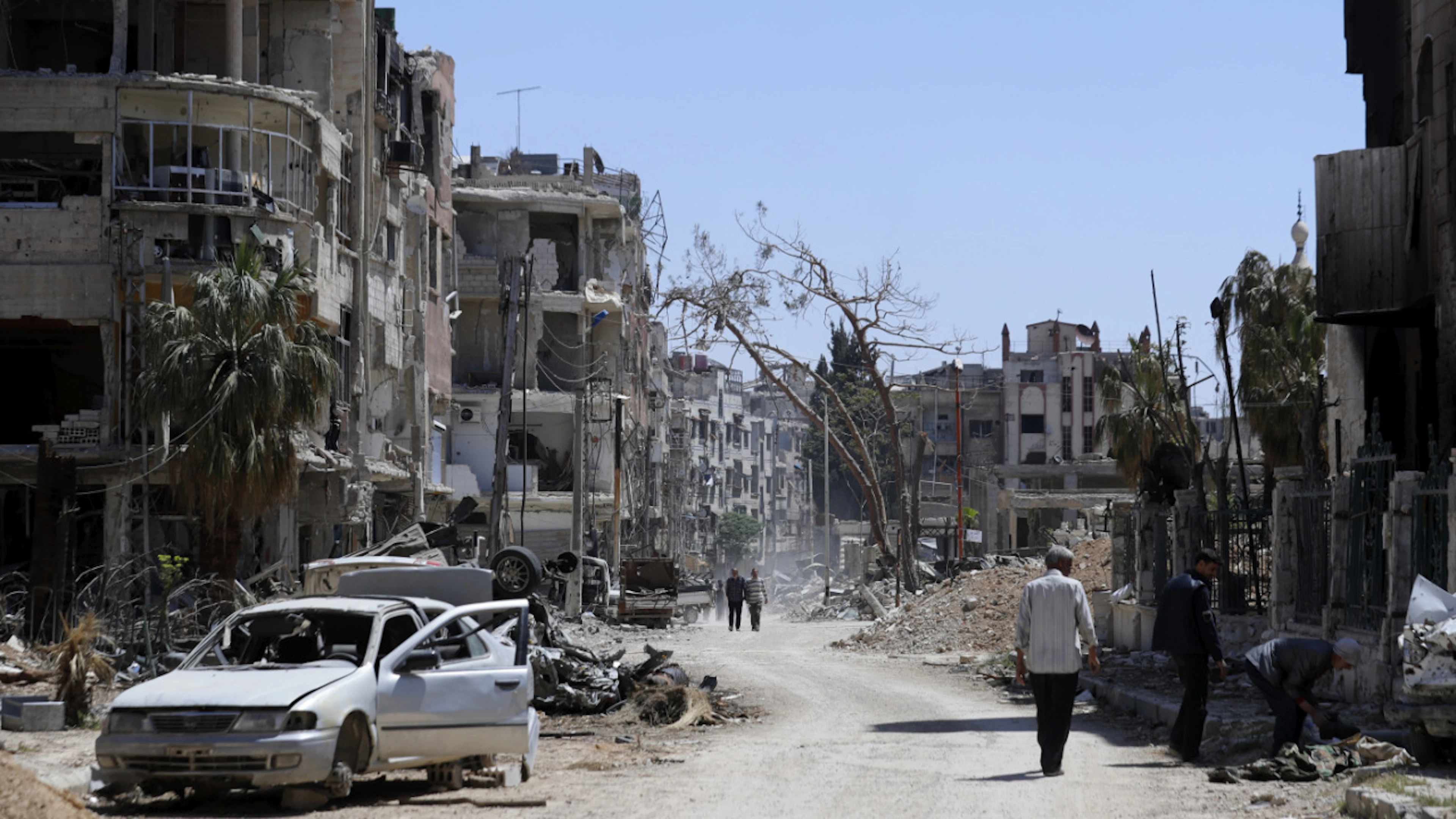 ARCHIV – 16.04.2018, Syrien, Duma: Menschen gehen an zerstörten Häusern und Autos im Vorort der syrischen Hauptstadt Damaskus vorbei. Foto: Hassan Ammar/AP/dpa