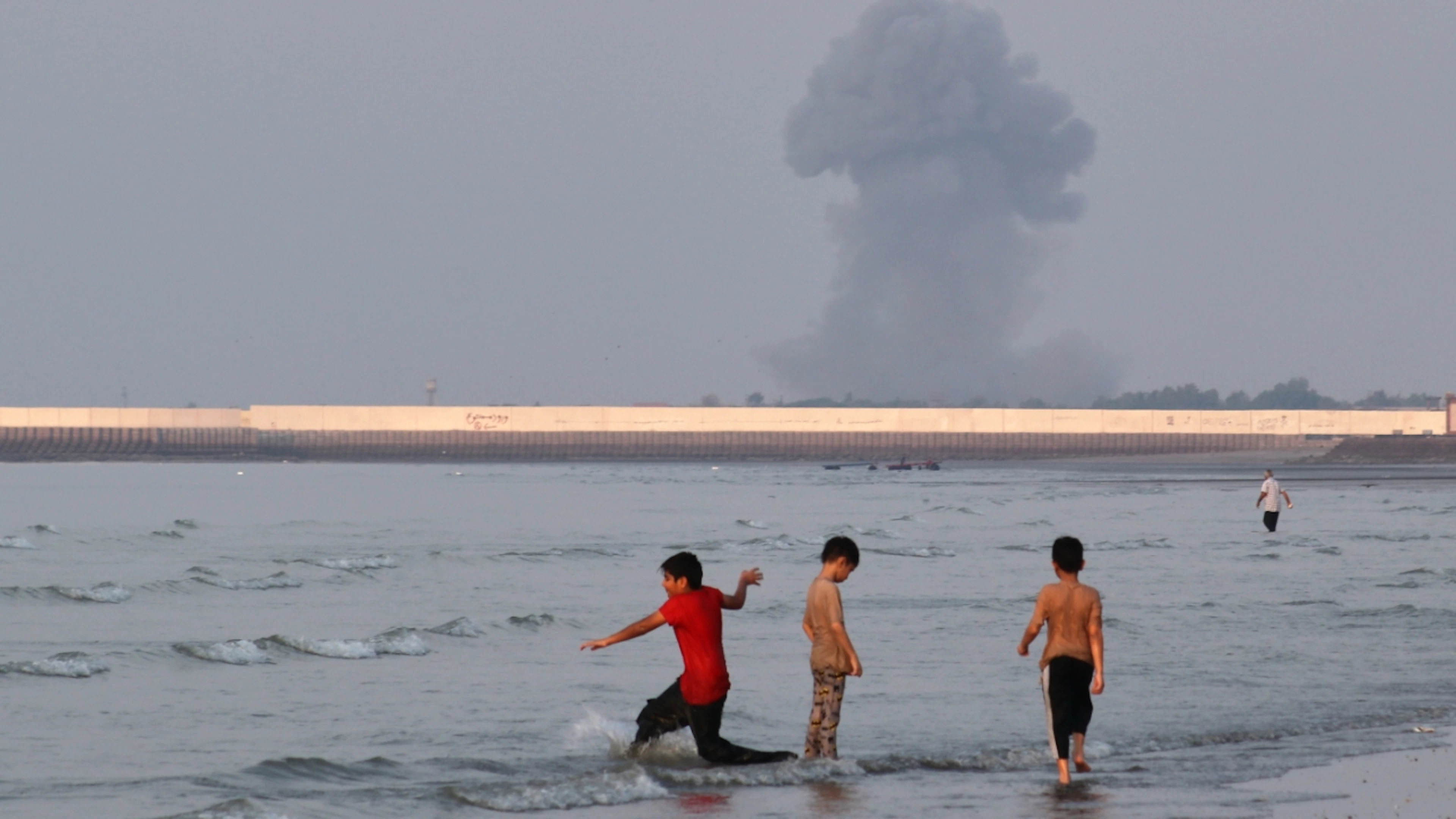 ARCHIVE – Three boys play in the shallow waters of the Strait of Hormuz, while in the background, off the coast of Bandar Abbas, Iran, a cloud of smoke rises following an explosion. Photo: Razieh Poudat/ISNA via AP/dpa
