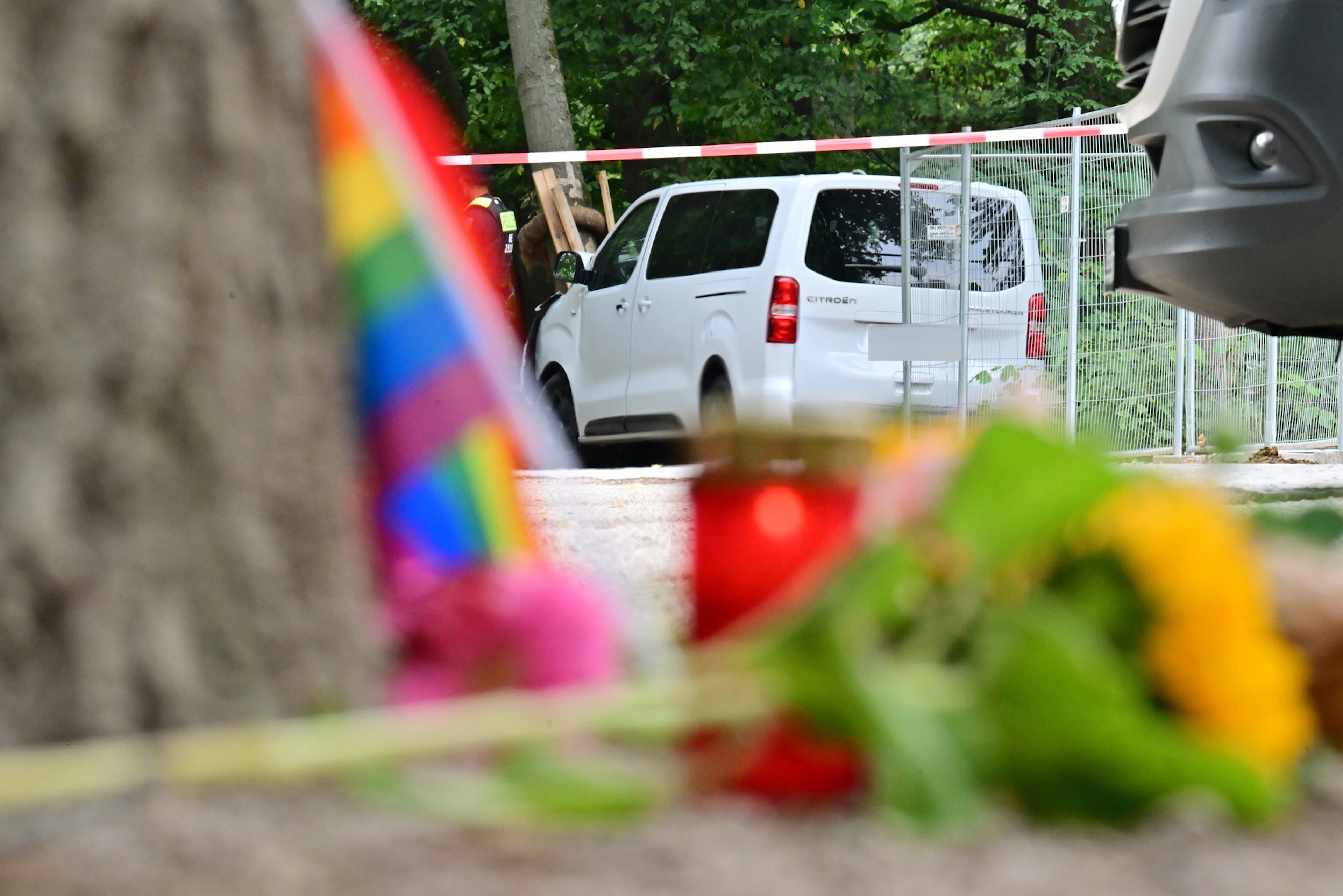 Fiori, candele e una bandiera arcobaleno davanti al veicolo bianco distrutto.